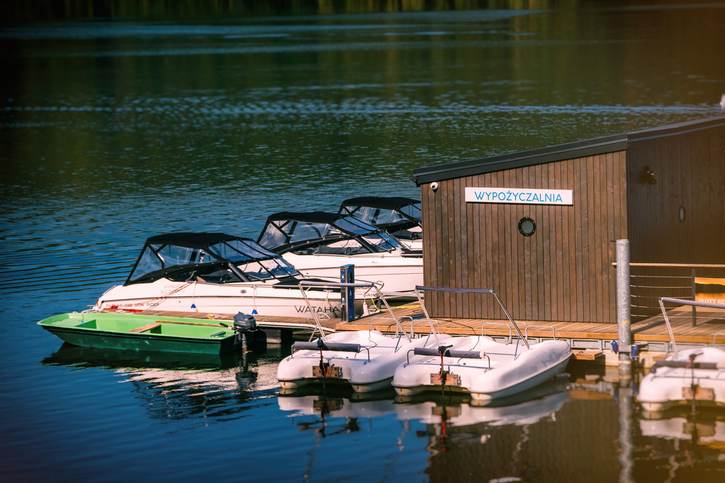 Część Jeziora Solińskiego, na jeziorze pomost, na pomoście drewniany, niewielki budynek z napisem „wypożyczalnia”. Do pomostu przycumowane białe rowerki wodne oraz biało-czarne, nowoczesne łodzie.