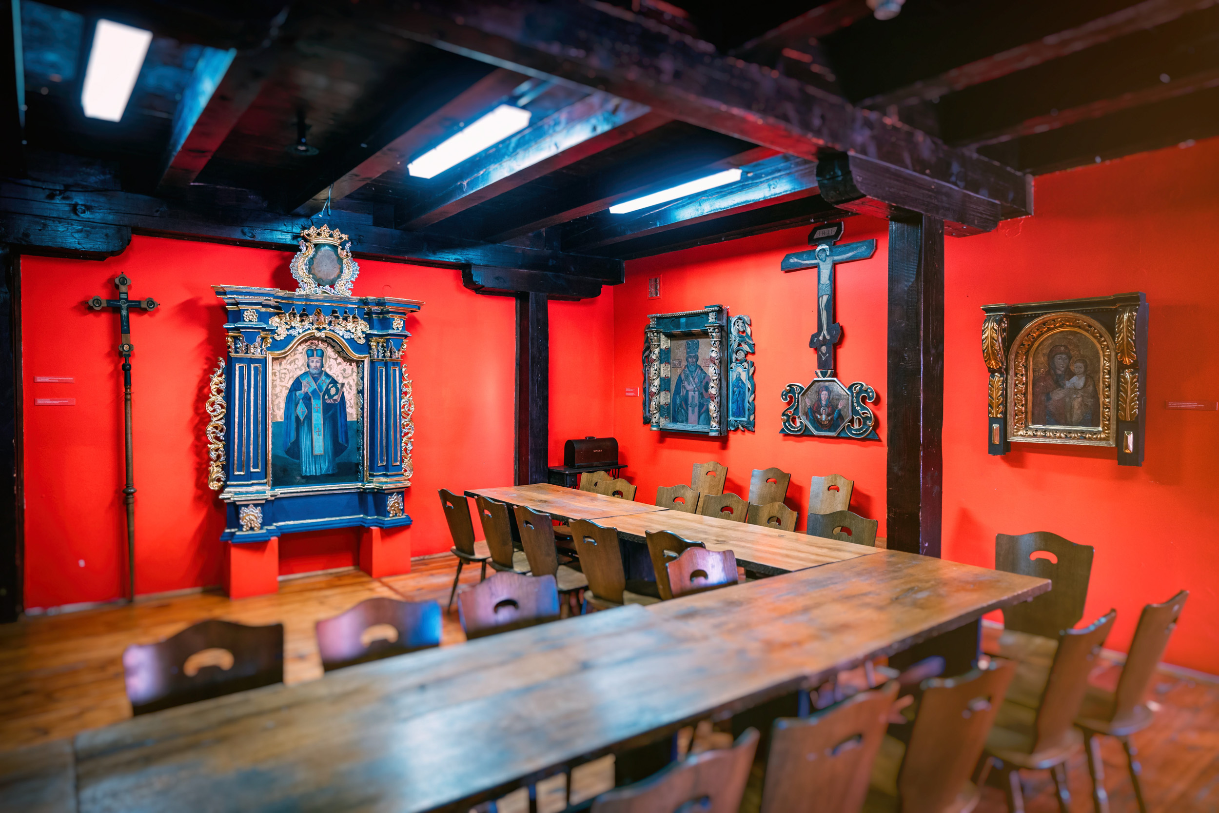 Na czerwonych ścianach zawieszone obrazy świętych, na środku sali drewniany stół z krzesłami, ciemnobrązowy drewniany sufit. 