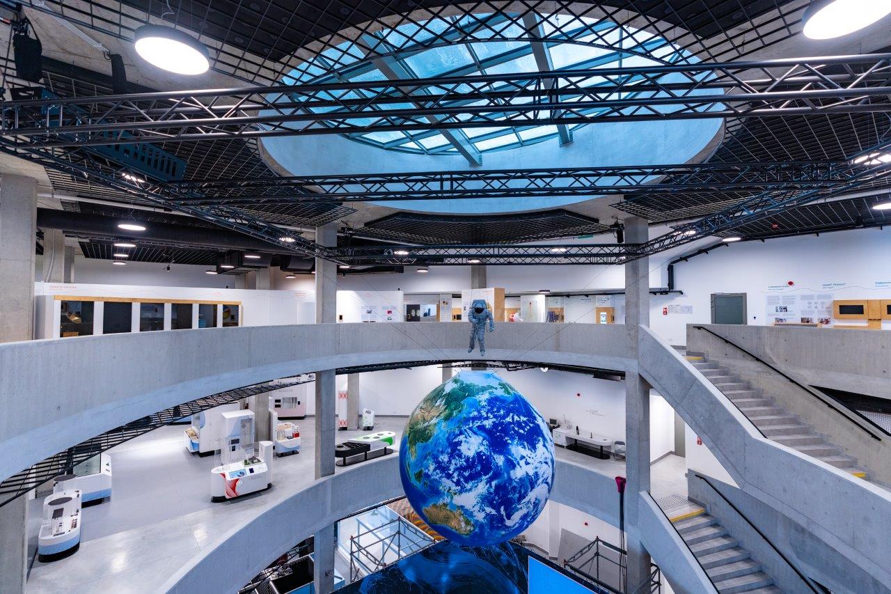 Atrium w trzykondygnacyjnym budynku widziane z góry. Schody wykonane z betonu, w tle słobo widoczne instalacje naukowe. Pod szklanym dachem zaiewszona ogromna makieta kuli ziemskiej obok niej kosmonauta.